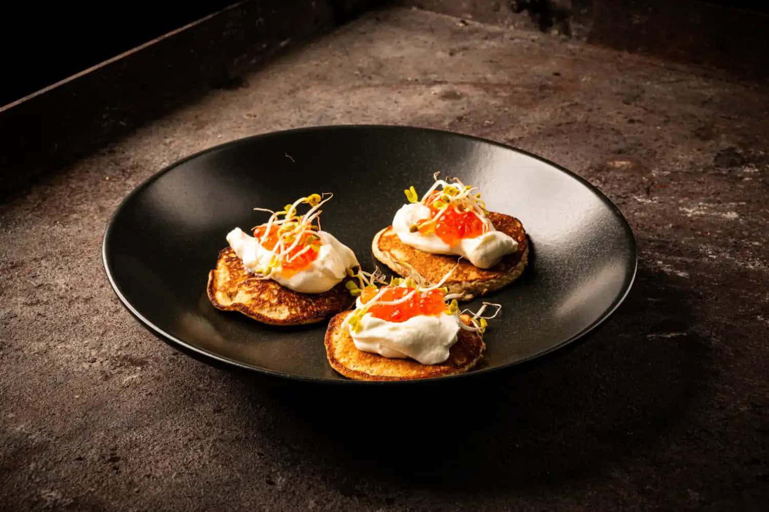 Blinis mit Frischkäse-Creme, geräuchertem Kaviar und Sprossen auf schwarzem Teller