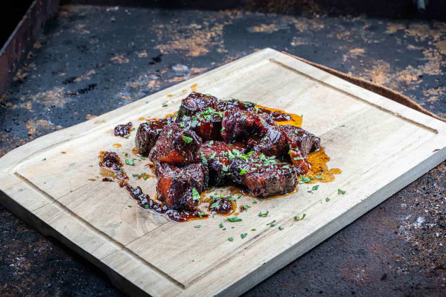 Glasierte Pork Belly Burnt Ends aus Schweinebauch-Würfeln in Barbecue-Sauce, garniert mit frischen Kräutern auf einem Schneidebrett.